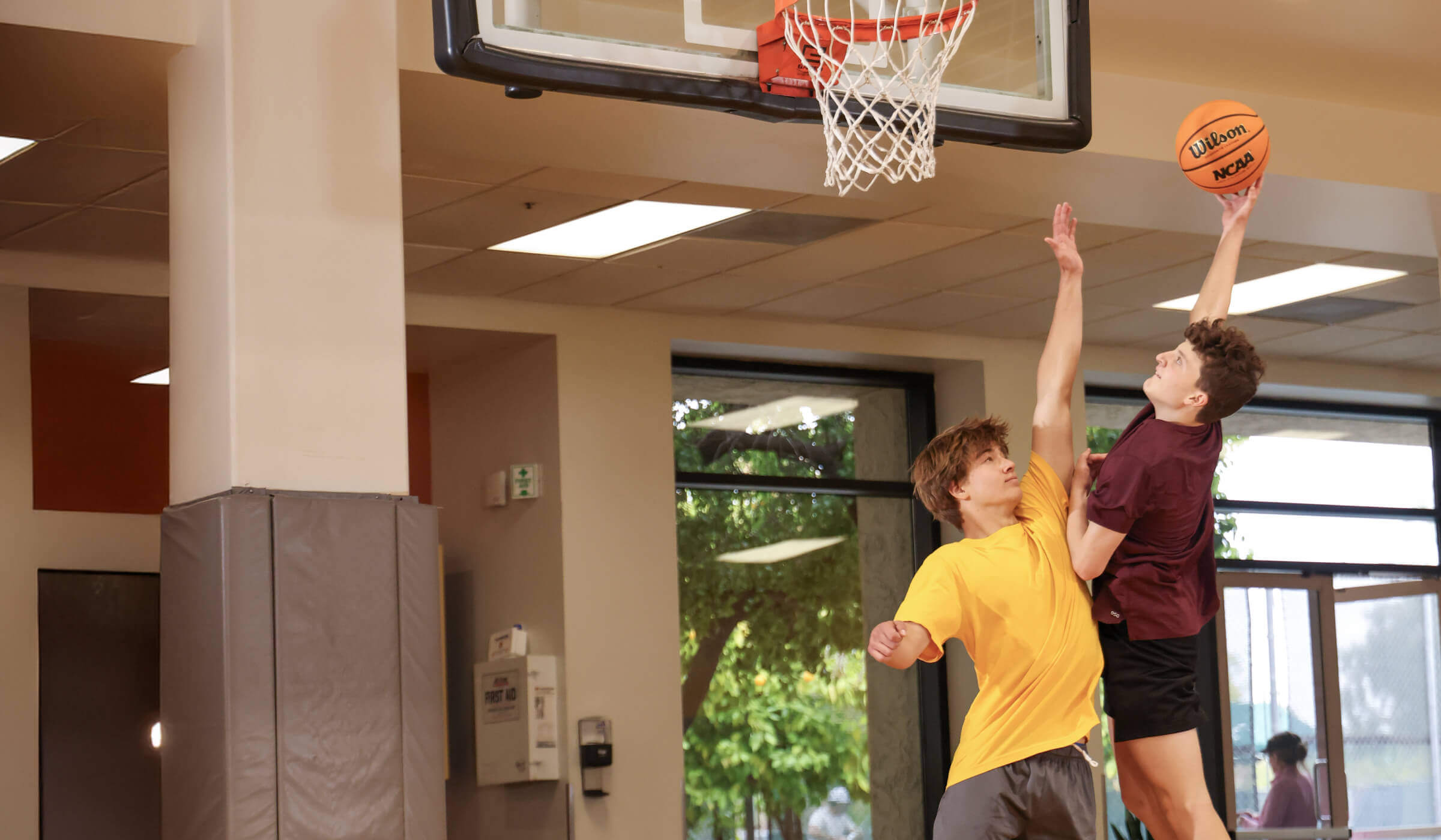 Two young people play basketball on an indoor court at Village Clubs Camelback, with greenery visible through large windows.