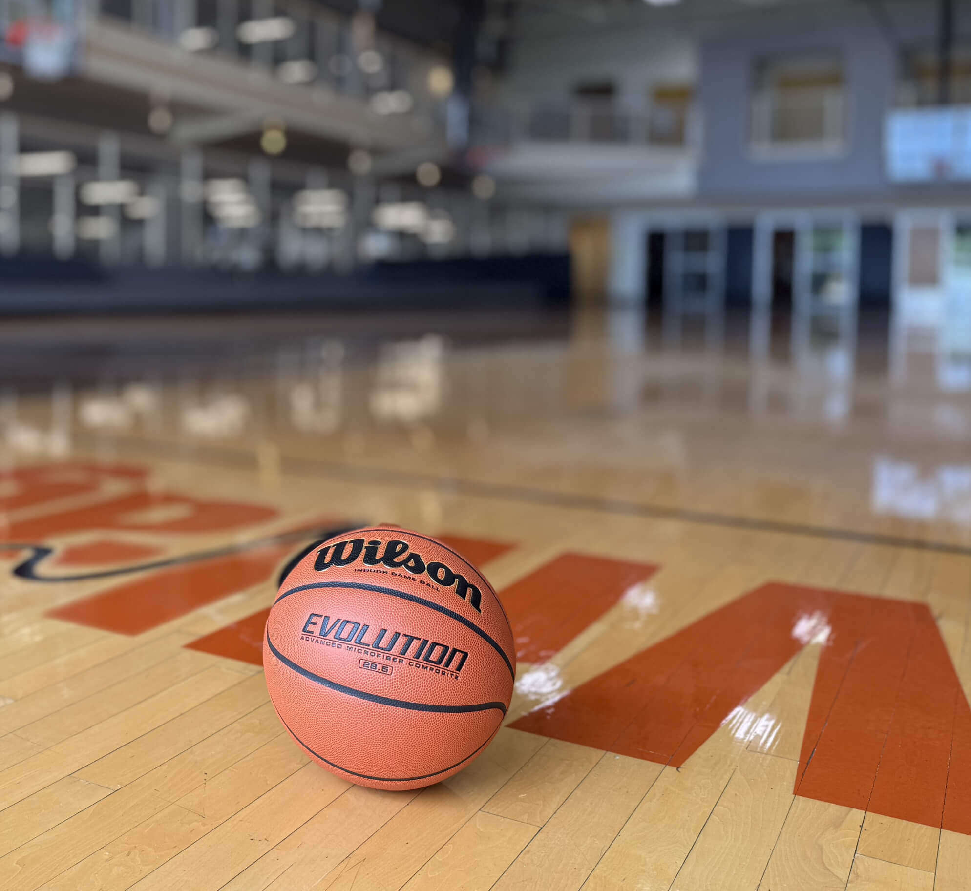 A Wilson Evolution basketball sits on the Village Clubs Camelback gym floor, with bright, empty courts and hoops in the background.