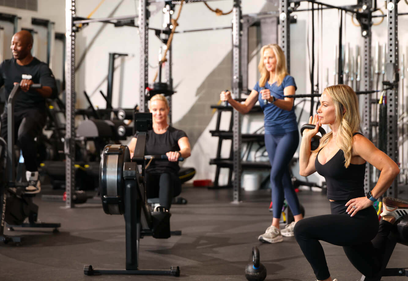 Members at Village Clubs Camelback work out on bikes, rowers, and weights in a spacious gym with strength racks behind them.