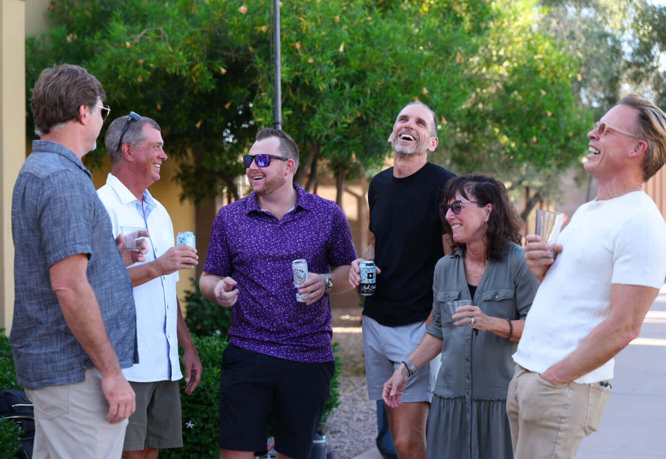 Adults enjoy a lively outdoor gathering at Village Clubs Camelback, socializing among greenery in a relaxed community setting.