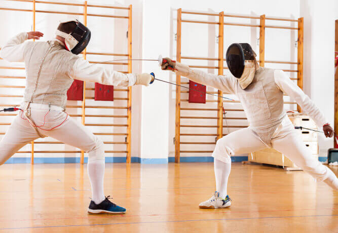 Two fencers in protective gear lunge with swords in the Village Clubs Camelback gym, featuring wood floors and wall bars.