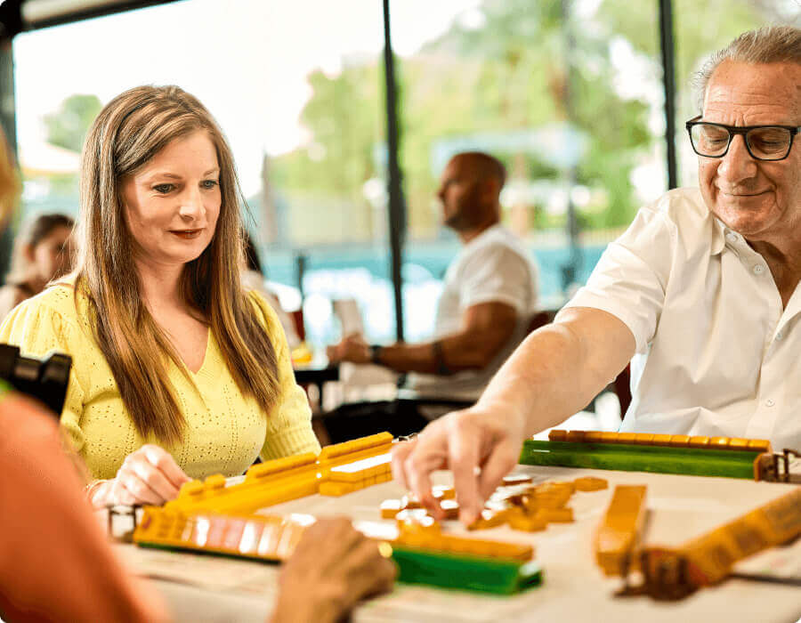 Members play a tile-based game at Village Clubs Camelback, focused at a table with others socializing in the background.
