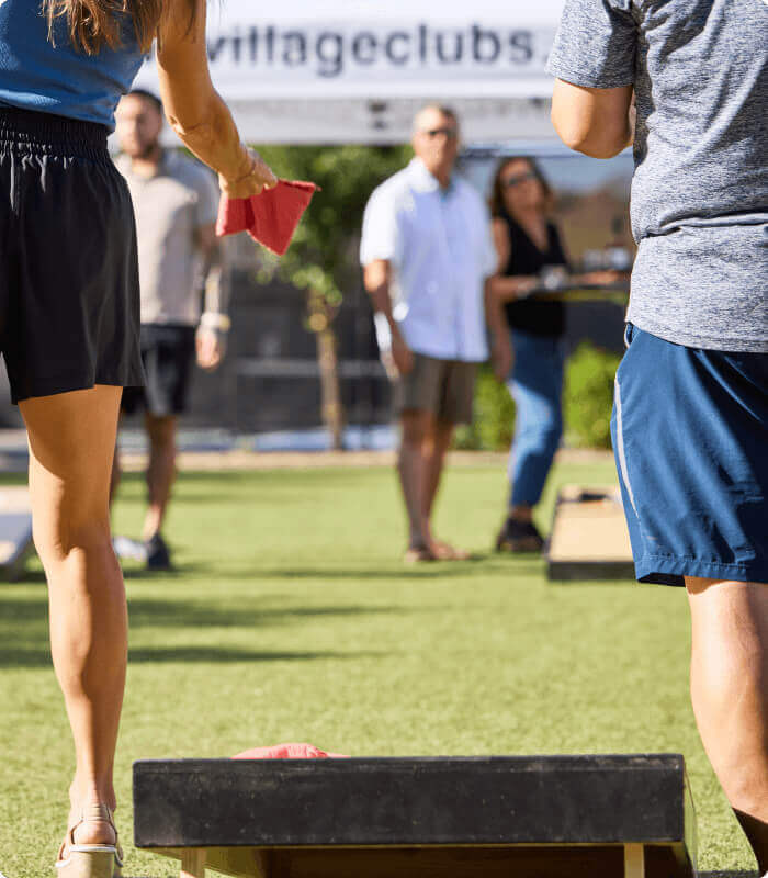 Members playing cornhole in the sun at Village Clubs Camelback, with others watching by the sign and grassy area.