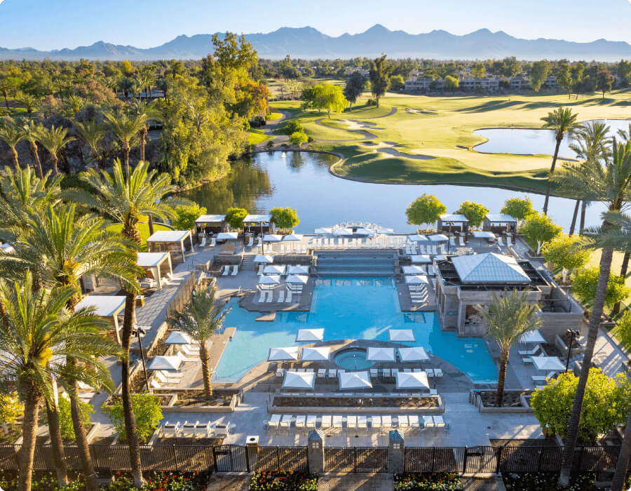 Aerial view of Village Clubs at Camelback featuring a large pool, sun loungers, cabanas, palm trees, and a golf course with mountain views.