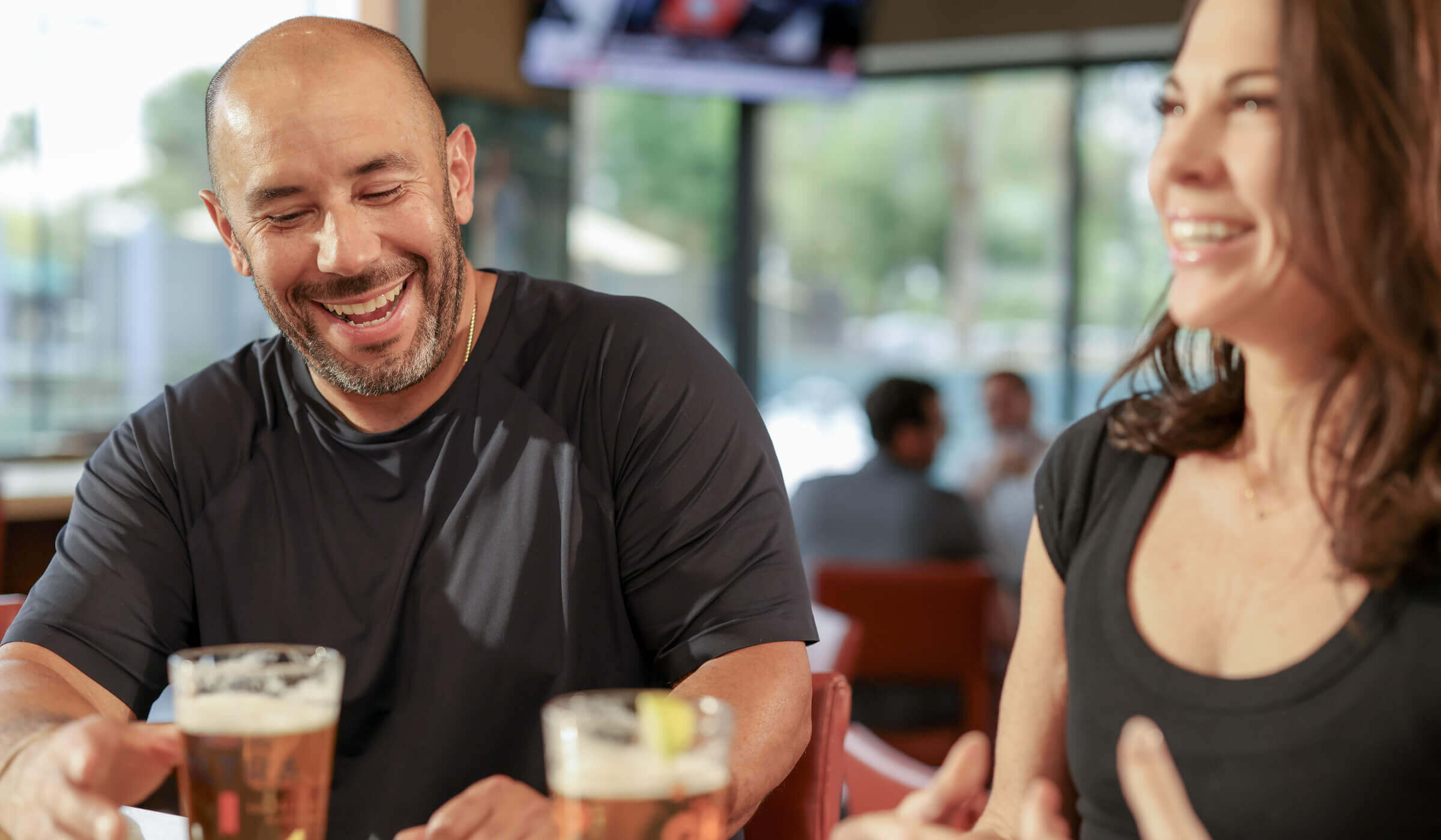 Two members enjoy drinks and laughter at Village Clubs Camelback, seated by sunlit windows in a vibrant dining area.