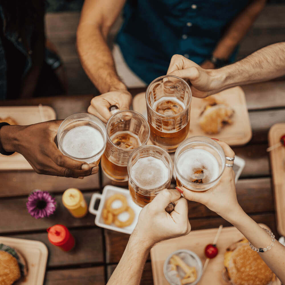 Five people toast beer mugs over burgers and fries at Village Clubs Camelback, highlighting membership perks and social dining.