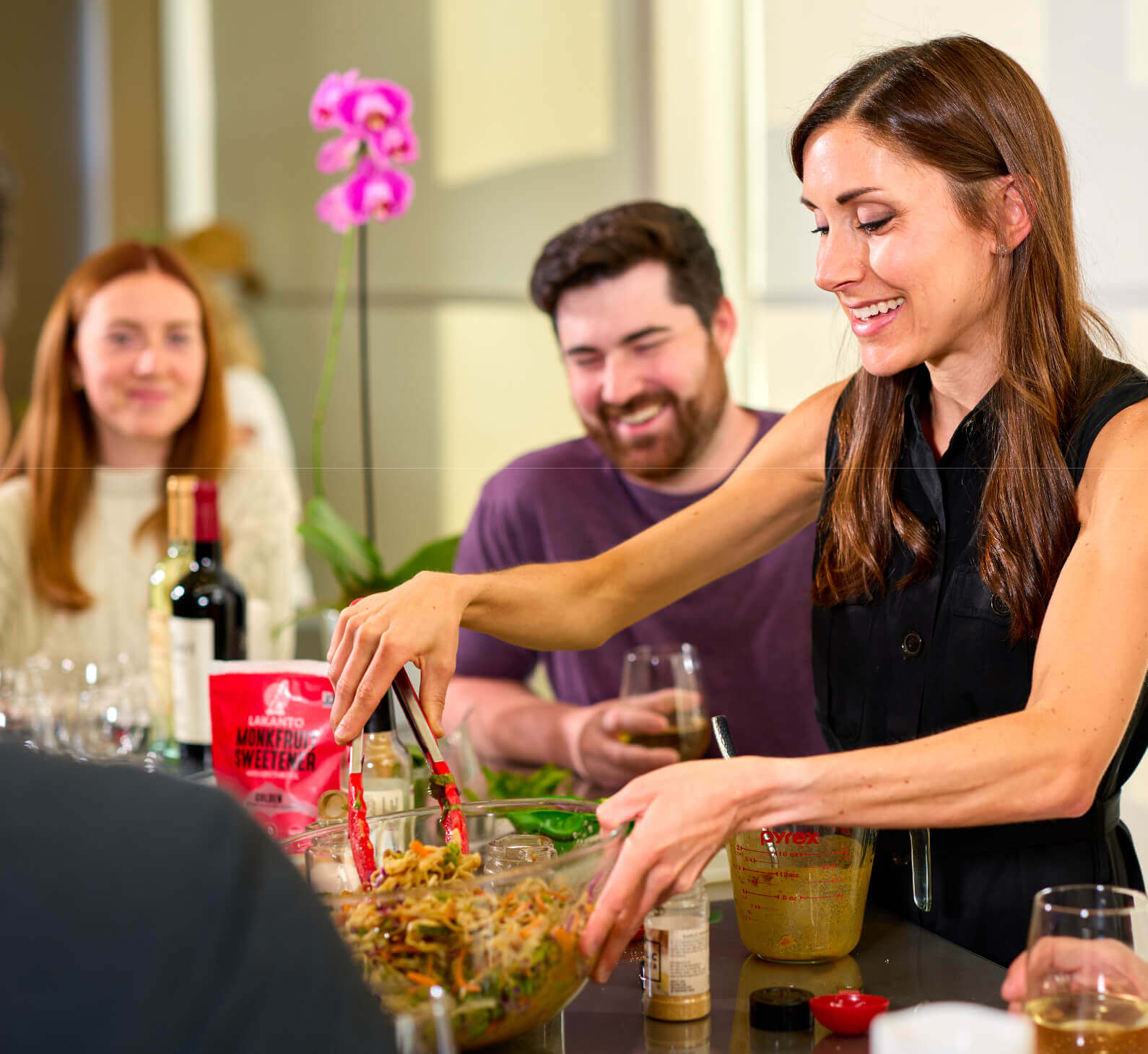 At Village Clubs Camelback, a woman serves food to smiling friends at a table set with drinks, ingredients, and a pink orchid.