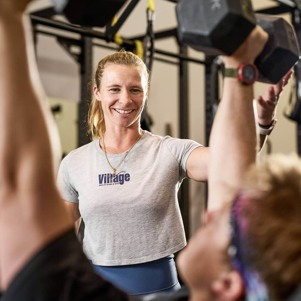 A Village Clubs Camelback trainer in a grey shirt supports a member lifting dumbbells overhead in the gym.