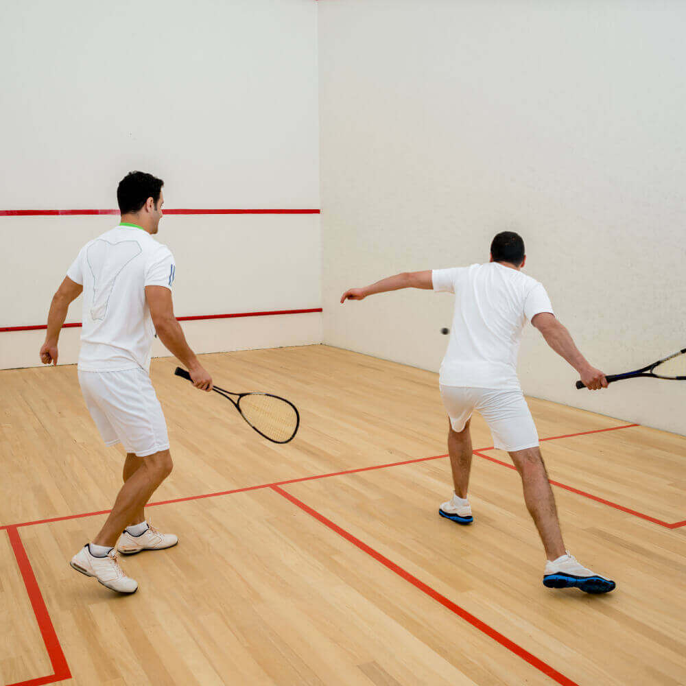 Two men play squash at Village Clubs Camelback, wearing white sportswear on a wooden court with red lines and squash rackets.