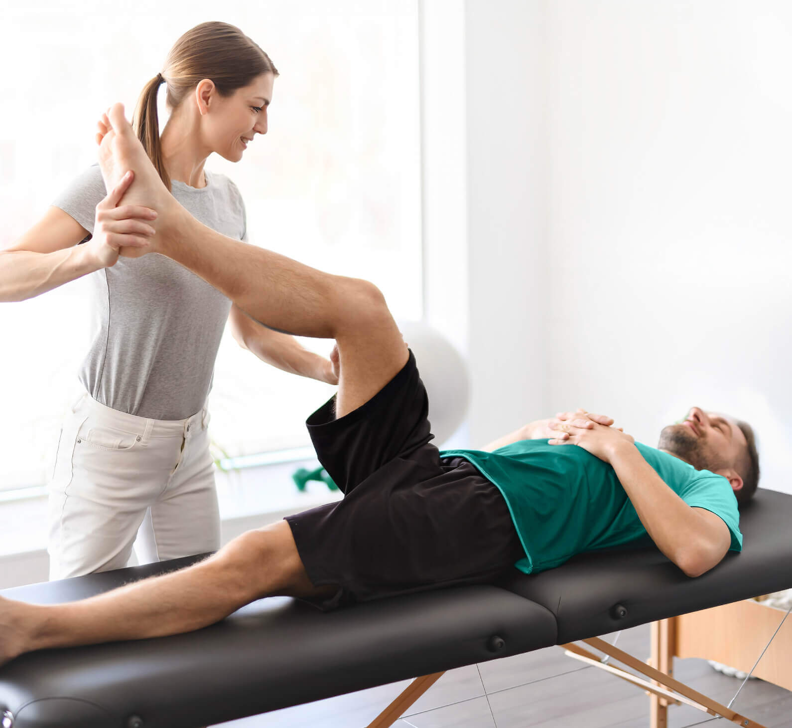 At Village Clubs Camelback, a physical therapist helps a man with leg stretches on a treatment table in a bright, modern clinic.