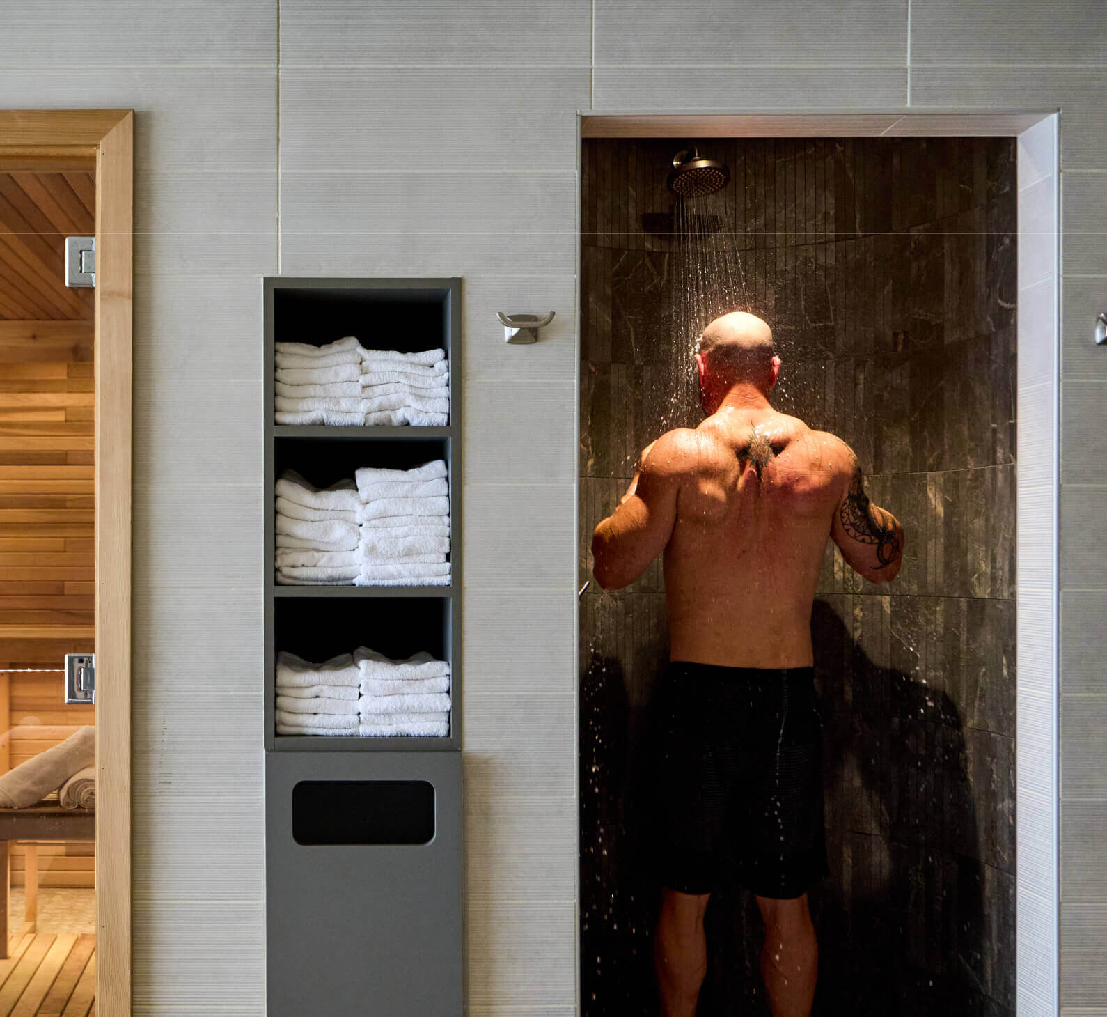 At Village Clubs Camelback, a man showers in a sleek, dark-tiled space with white towels and sauna therapy visible nearby.