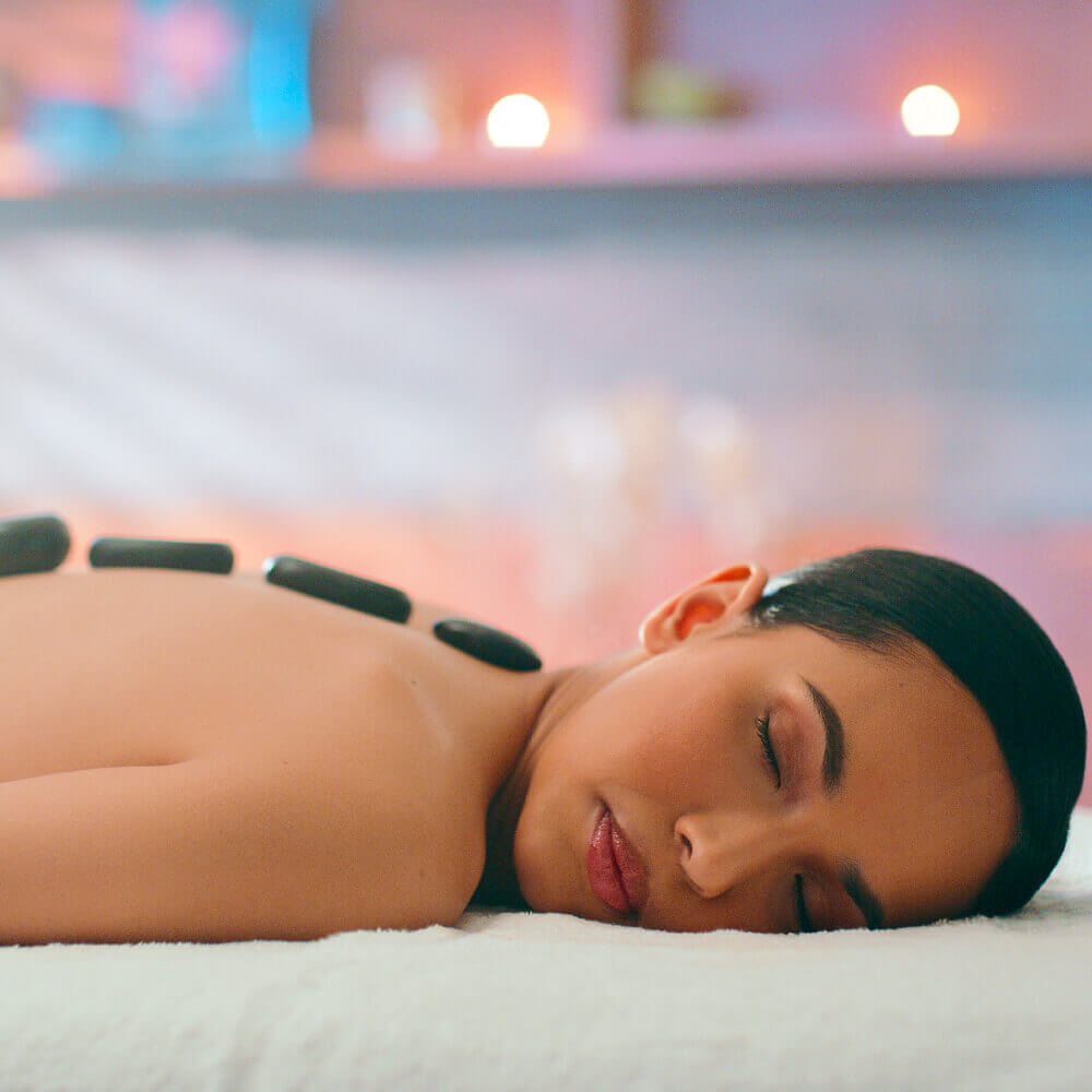 A woman enjoys hot stone therapy at Village Clubs Camelback spa, relaxing on a massage table in a softly lit, calm setting.