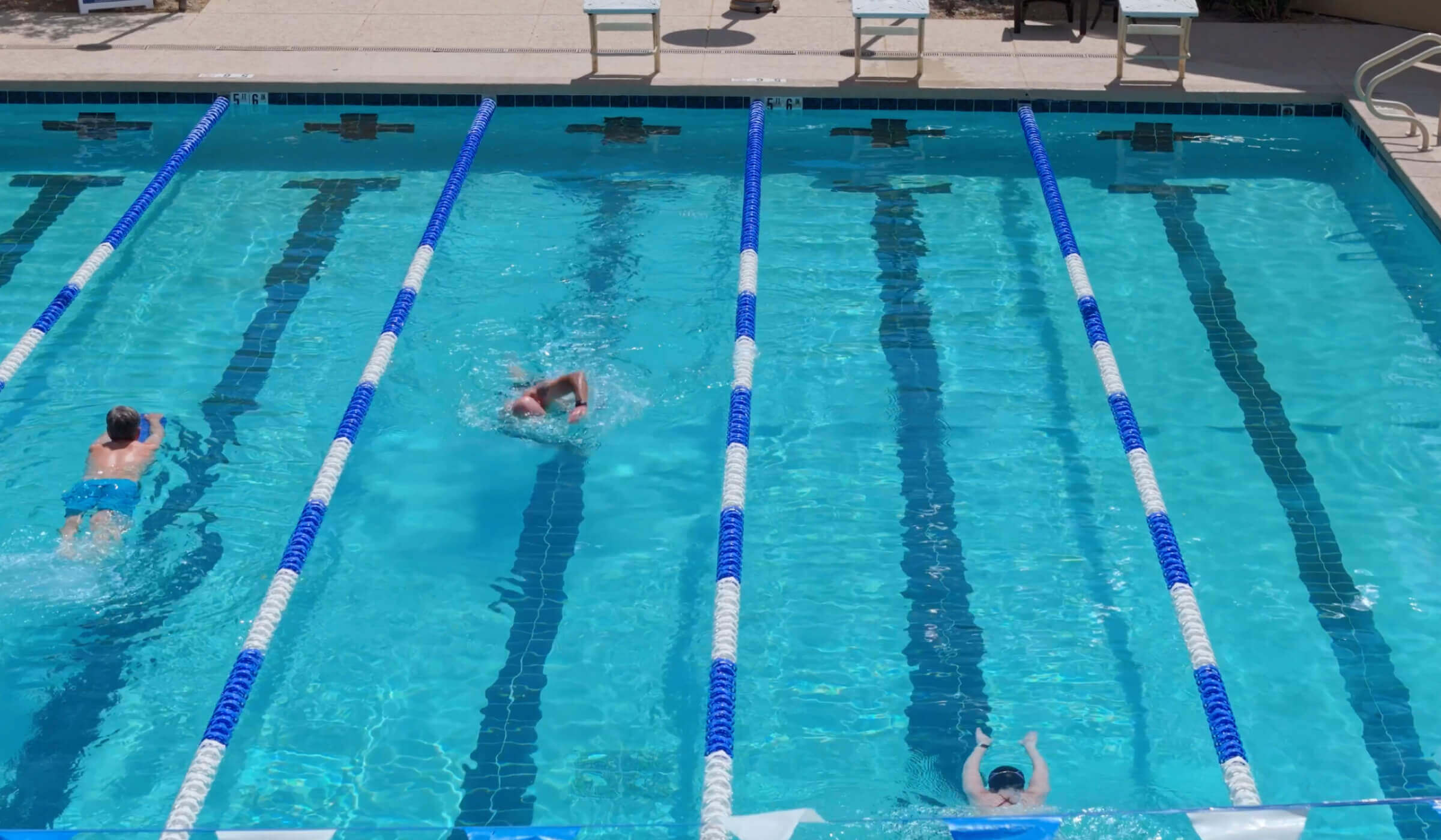 Swimmers compete in outdoor lanes at Village Clubs Camelback, Arizona, with blue dividers and clear water visible from above.