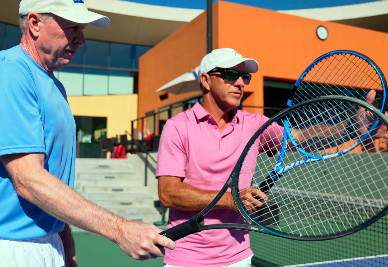 Two men discuss tennis rackets on a Village Clubs court at Camelback, with a modern clubhouse and stairs in the background.