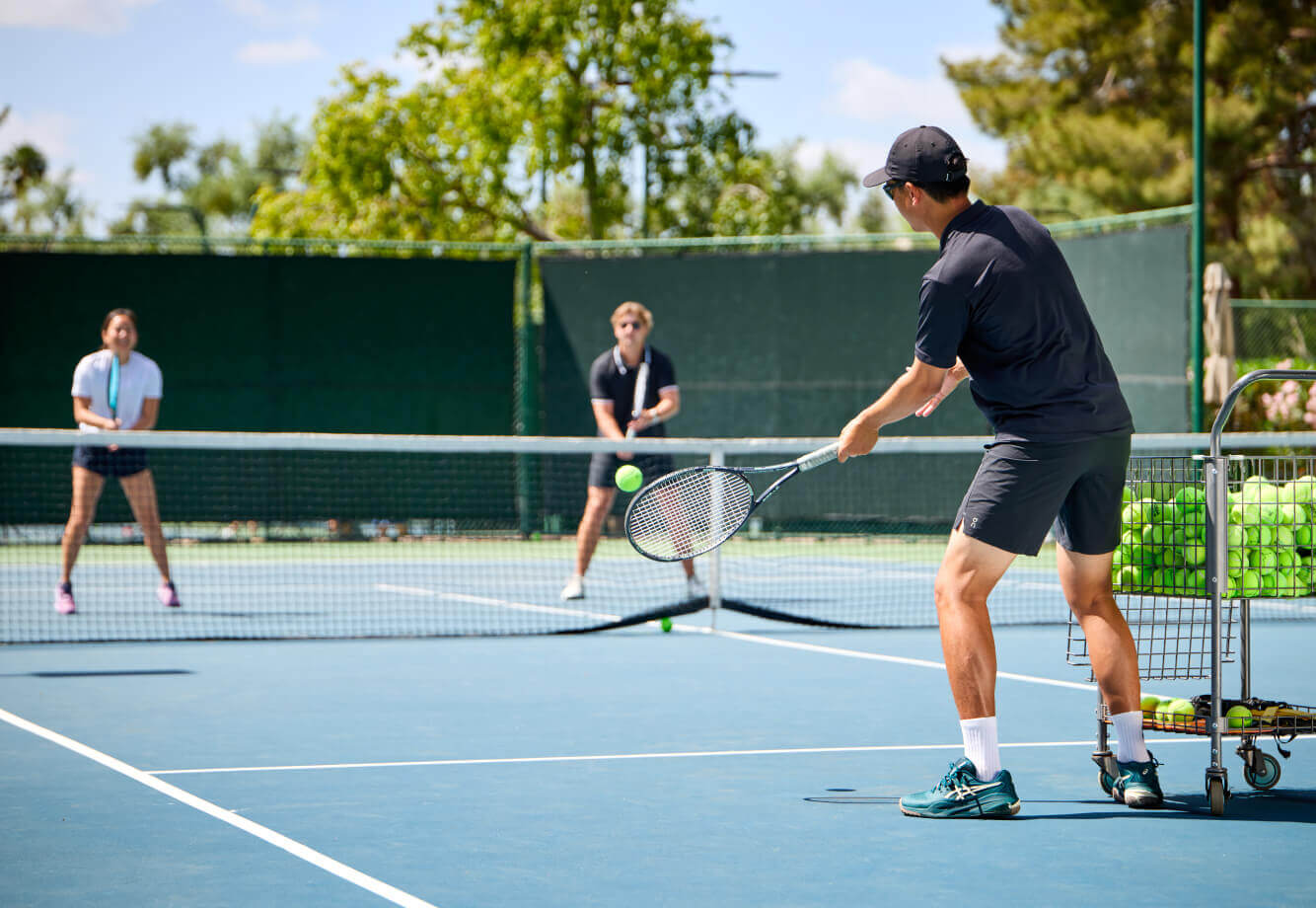 Village Clubs tennis lesson at DC Ranch: coach practices with two students on a blue court; basket of balls nearby in sunny weather.