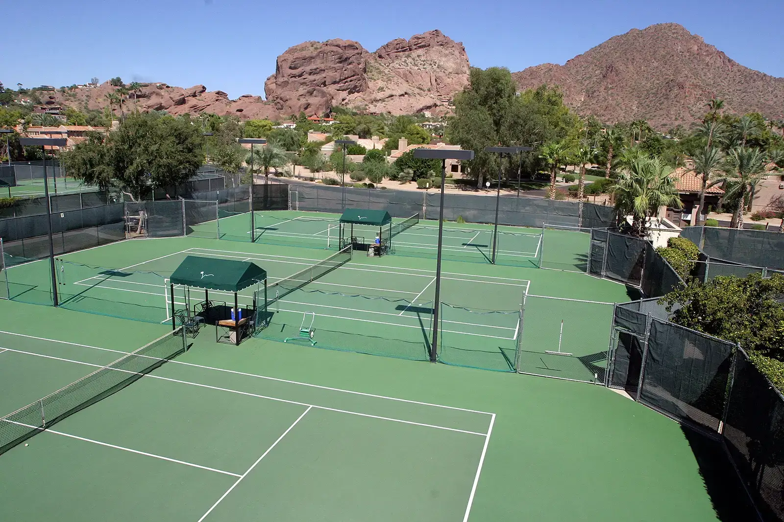 Village Clubs Camelback outdoor tennis courts with nets, shaded benches, palm trees, and mountain views under a clear blue sky.