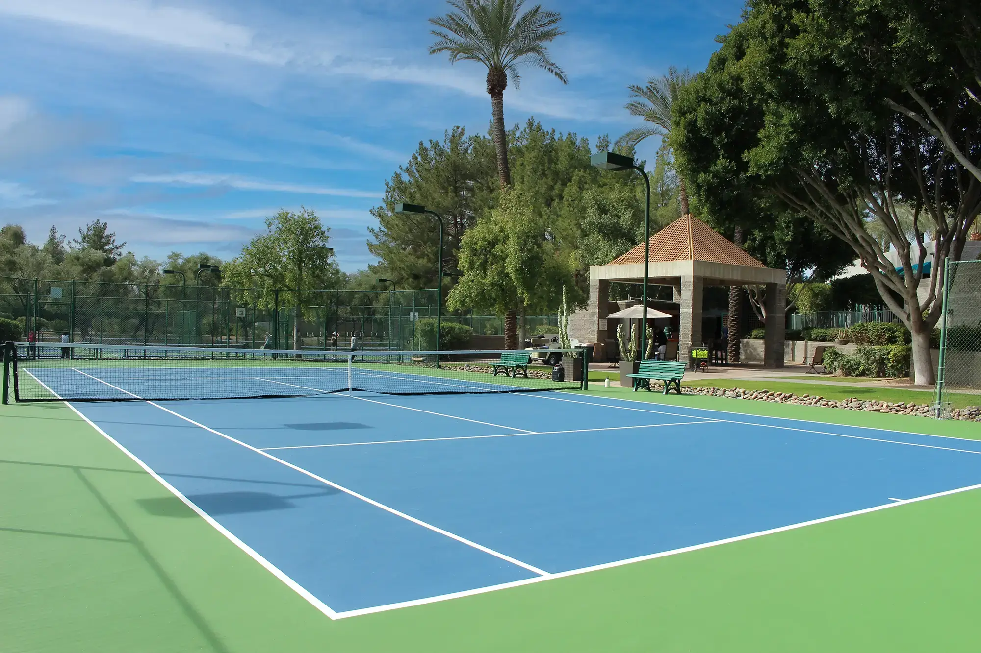Village Clubs Camelback outdoor tennis courts with blue surfaces, green borders, pavilion, and benches amid lush Arizona greenery.