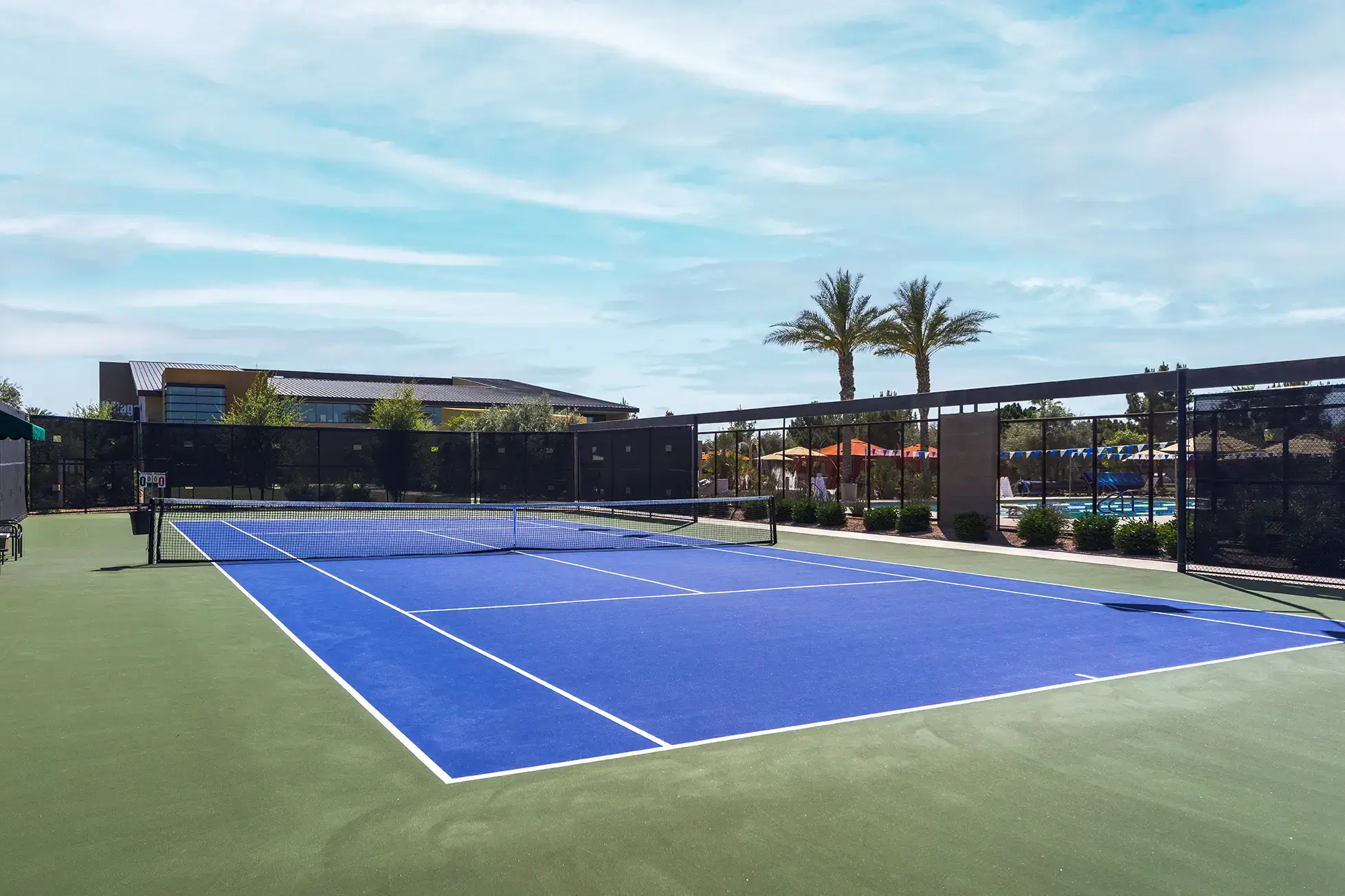 Village Clubs Camelback outdoor tennis court with blue and green surface, palm trees, and modern buildings under a partly cloudy sky.