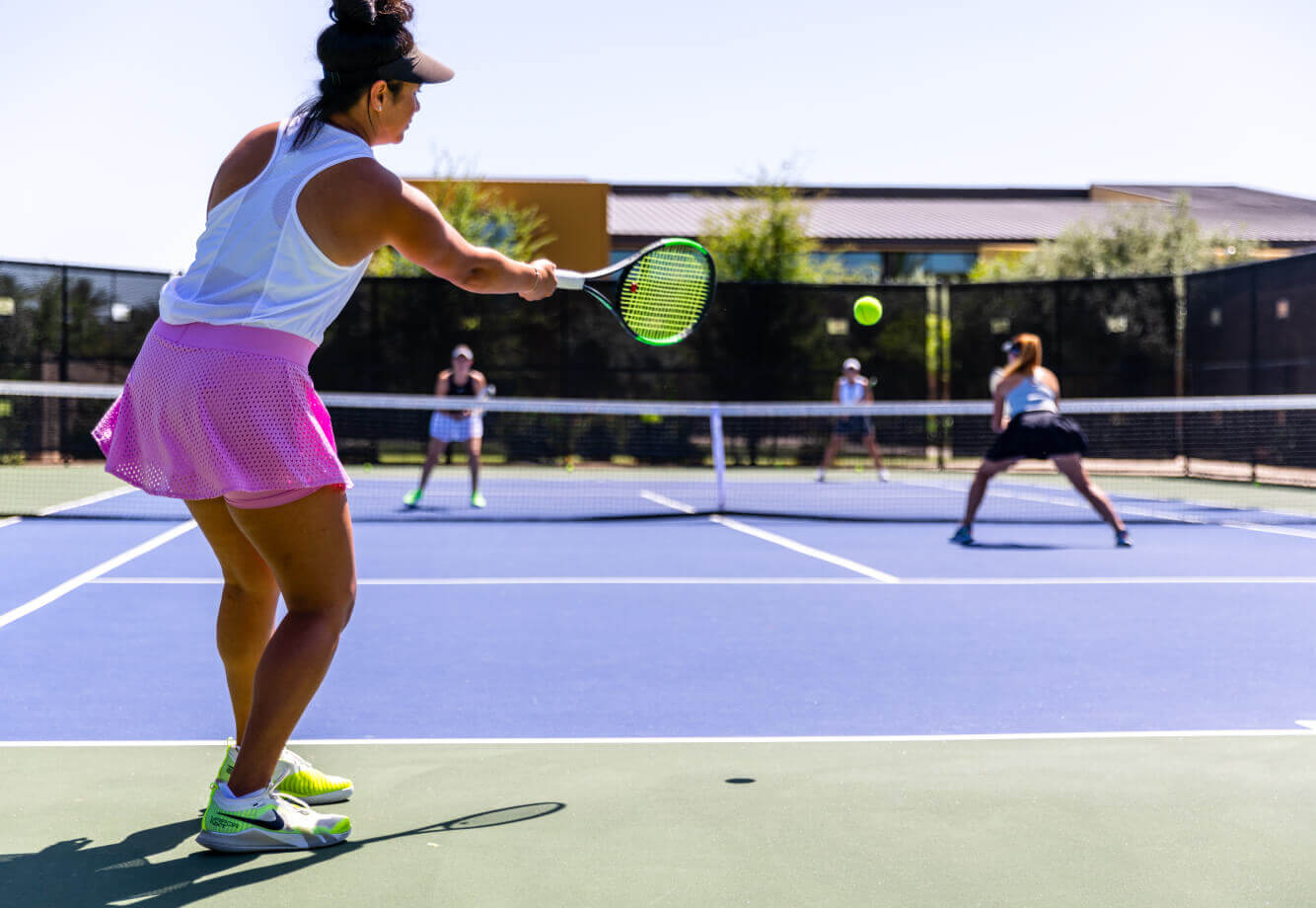At Village Clubs DC Ranch, women play tennis on a sunlit outdoor court, with one serving and others ready to return.