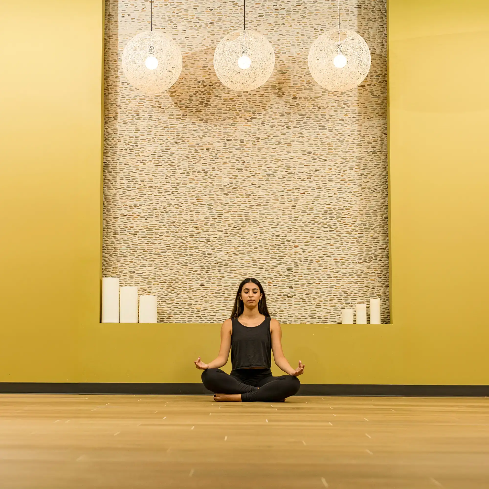 A woman meditates cross-legged on a wooden floor in a serene Village Clubs Camelback studio with yellow walls and soft lighting.