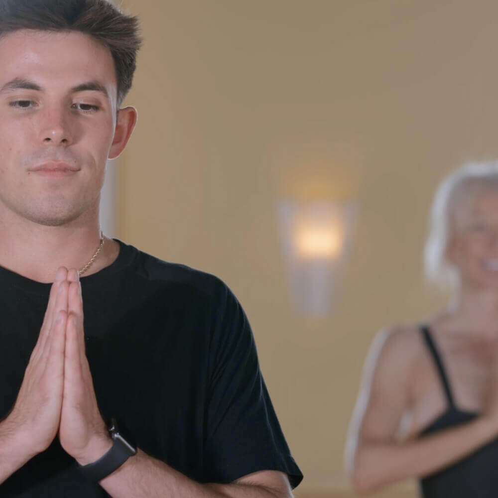 A young man practices yoga at Village Clubs Camelback, hands in prayer position, eyes closed; another member poses behind him.