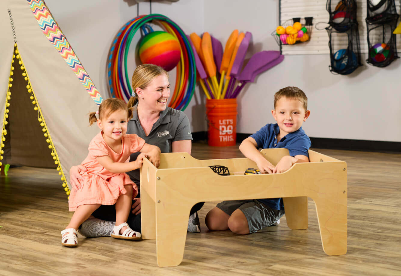 A woman and two kids play at a wooden activity table in Village Clubs Camelback’s colorful indoor playroom with sports toys nearby.